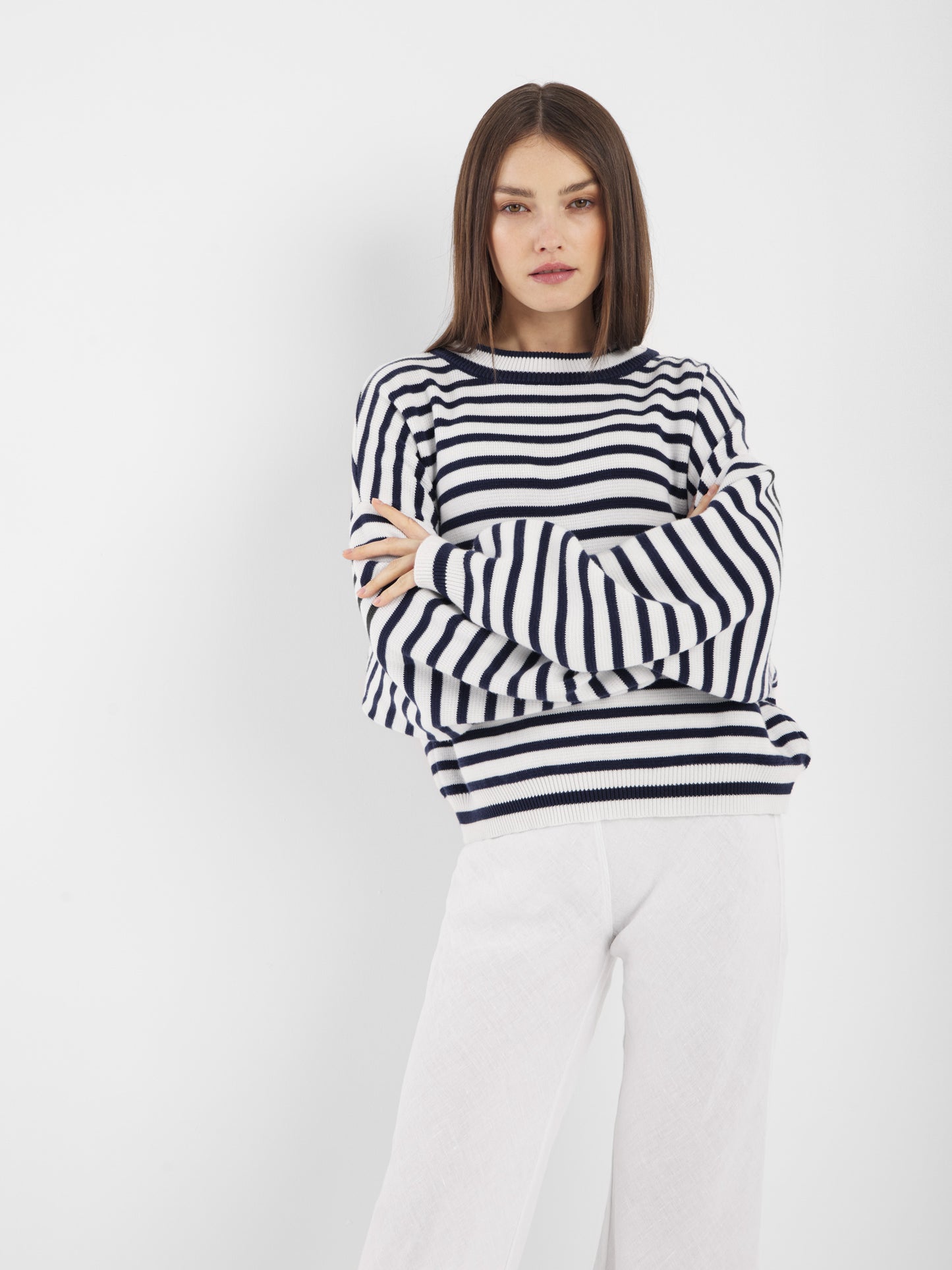 Eine Frau mit glattem braunem Haar steht vor einem weißen Hintergrund und trägt den Pullover Sailor Navy White und eine weiße Hose. Ihre Arme sind verschränkt und sie hat einen neutralen Gesichtsausdruck.