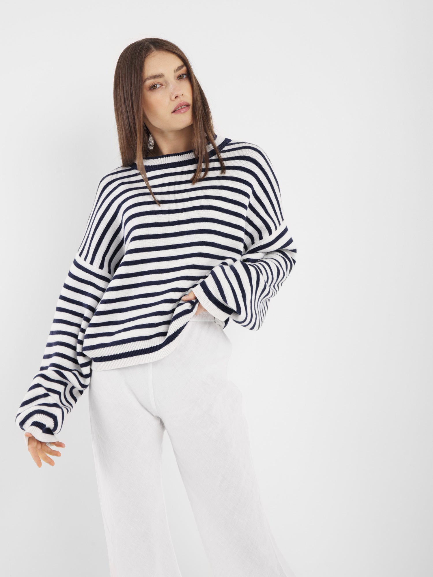 Eine Person mit glattem braunem Haar trägt den Pullover Sailor Navy White und eine weitbeinige weiße Hose vor einem schlichten weißen Hintergrund.