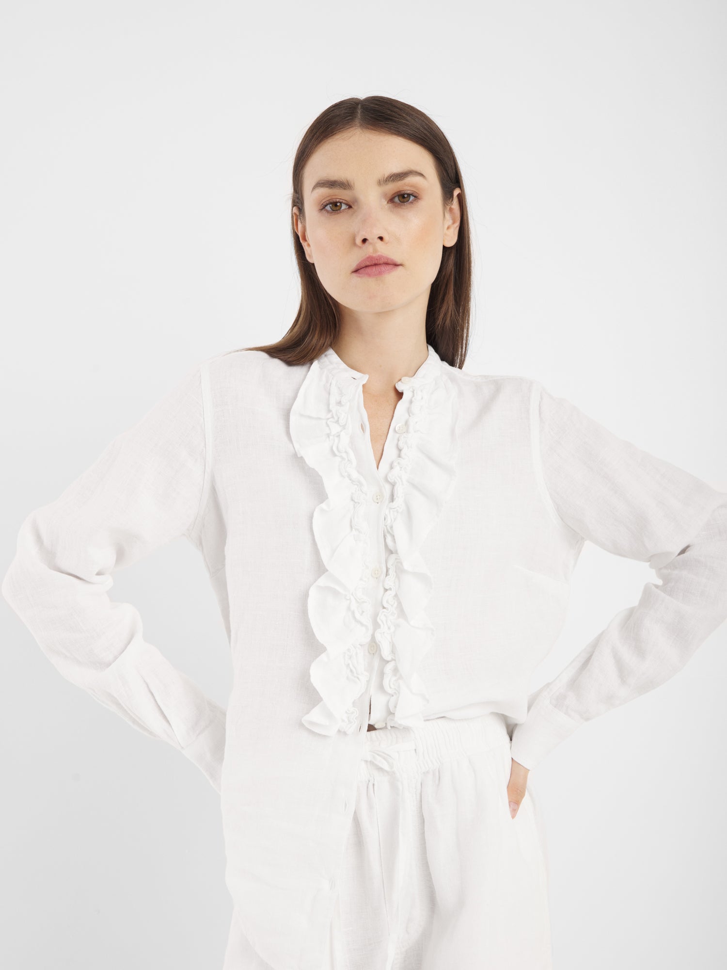 Eine Frau mit glattem braunem Haar modelliert das Bluse Ruffle Linen White mit passender weißer Hose und steht selbstbewusst vor einem schlichten weißen Hintergrund, die Hände in die Hüften gestemmt.
