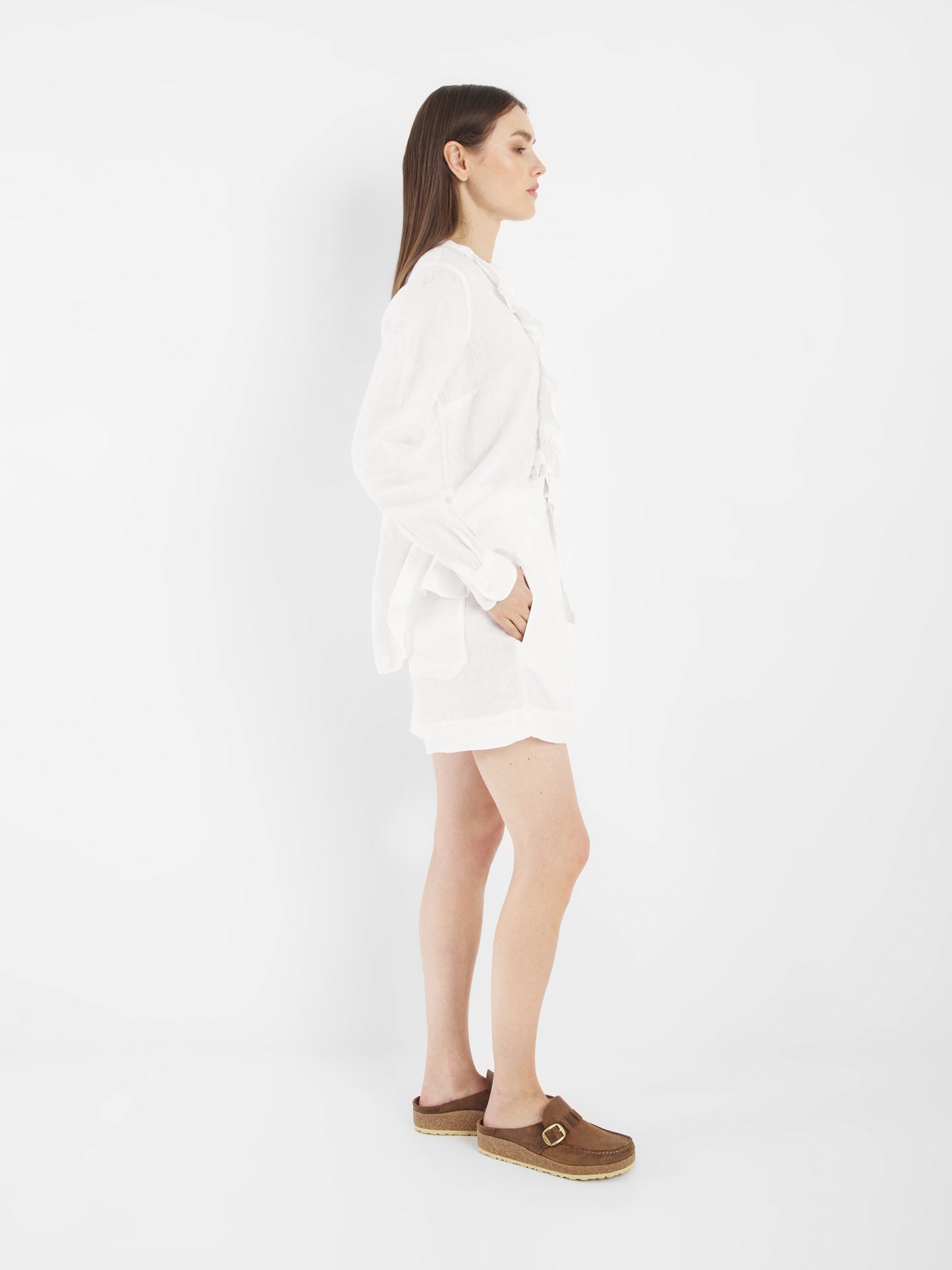 Eine Frau steht im Profil vor einem weißen Hintergrund. Sie trägt die Bluse Ruffle Linen White mit langen Ärmeln, kombiniert mit weißen Shorts und braunen Slippern mit Schnallen. Ihre linke Hand steckt in ihrer Tasche und ihr Haar ist glatt und offen.