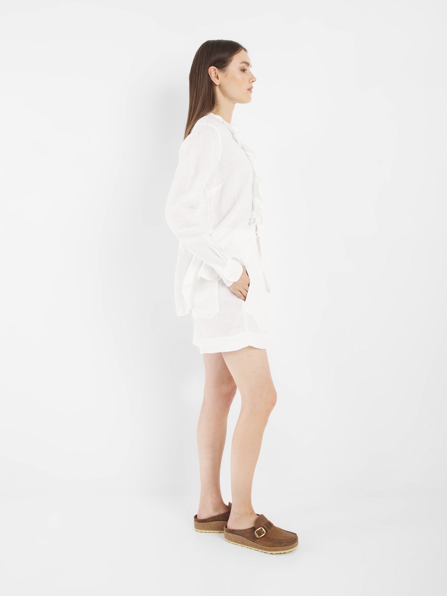 Eine Frau steht im Profil vor einem weißen Hintergrund. Sie trägt die Bluse Ruffle Linen White mit langen Ärmeln, kombiniert mit weißen Shorts und braunen Slippern mit Schnallen. Ihre linke Hand steckt in ihrer Tasche und ihr Haar ist glatt und offen.
