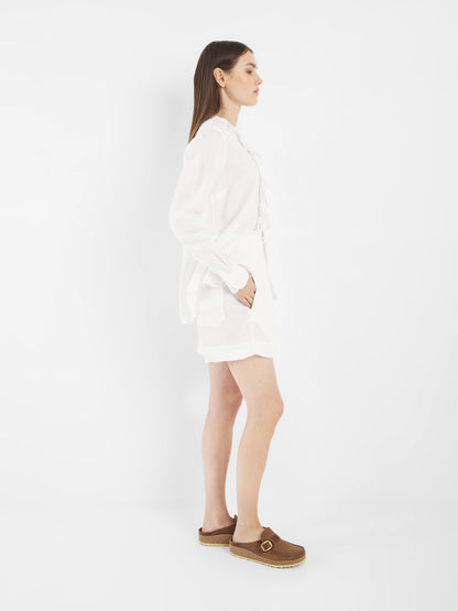 Eine Frau steht im Profil vor einem weißen Hintergrund. Sie trägt die Bluse Ruffle Linen White mit langen Ärmeln, kombiniert mit weißen Shorts und braunen Slippern mit Schnallen. Ihre linke Hand steckt in ihrer Tasche und ihr Haar ist glatt und offen.