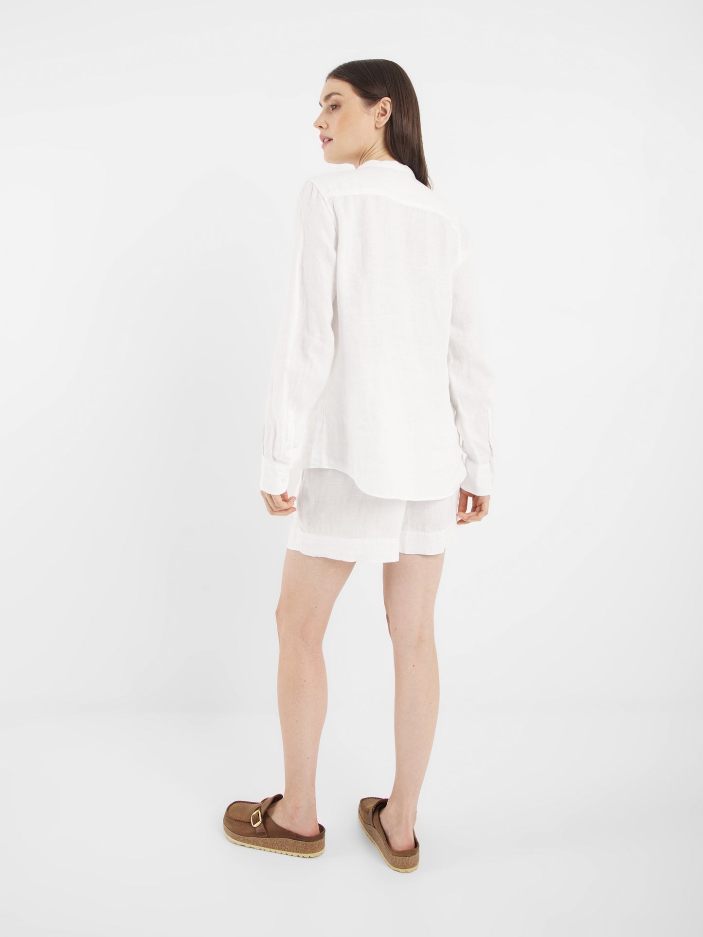 Eine Frau blickt von sich weg und leicht zur Seite. Sie trägt ein langärmeliges Bluse Ruffle Linen White, weiße Shorts und braune Slipper-Sandalen vor einem schlichten weißen Hintergrund.