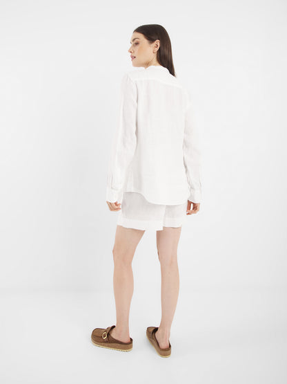 Eine Frau blickt von sich weg und leicht zur Seite. Sie trägt ein langärmeliges Bluse Ruffle Linen White, weiße Shorts und braune Slipper-Sandalen vor einem schlichten weißen Hintergrund.