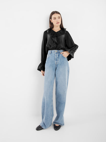 Eine Frau mit glattem braunem Haar und neutralem Gesichtsausdruck steht vor einem weißen Hintergrund. Sie trägt das Oberteil Ruffle Long Sleeve Black, hellblaue Jeans mit hoher Taille und weitem Bein sowie schwarze Schuhe.