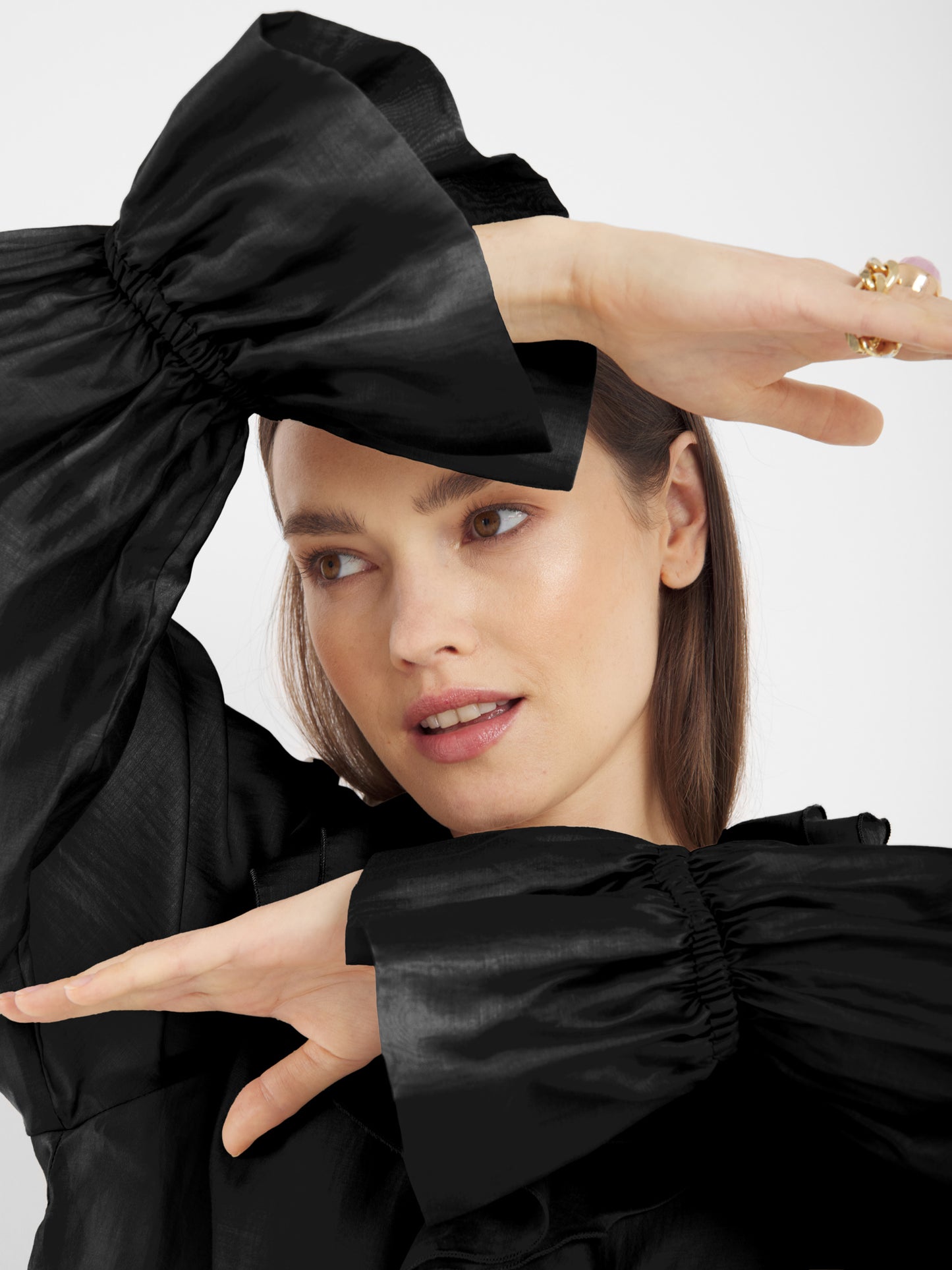 Eine Frau mit heller Haut und glattem braunem Haar posiert mit erhobenen Armen und trägt das Ruffle Long Sleeve Black - ein elegantes Beispiel für nachhaltige Mode. Sie blickt sanft zur Seite.
