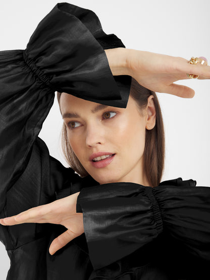 Eine Frau mit heller Haut und glattem braunem Haar posiert mit erhobenen Armen und trägt das Ruffle Long Sleeve Black - ein elegantes Beispiel für nachhaltige Mode. Sie blickt sanft zur Seite.