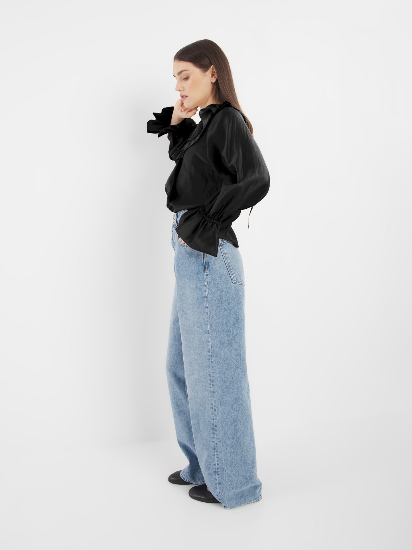 Eine Frau mit langen braunen Haaren steht seitlich vor einem weißen Hintergrund. Sie trägt die Ruffle Long Sleeve Black Bluse und hellblaue Jeans mit weiten Beinen - ein eleganter Look für nachhaltige Mode, und eine Hand berührt ihr Gesicht, während sie nach unten schaut.