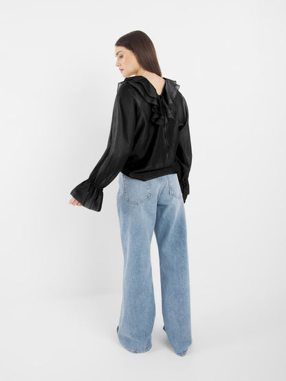 Eine Frau mit langen braunen Haaren steht mit dem Gesicht zur Wand. Sie trägt die schwarze Bluse Ruffle Long Sleeve Black mit Rüschendetails und weiten Ärmeln, gepaart mit hellblauen Jeans mit hoher Taille und weitem Bein vor einem schlichten weißen Hintergrund.