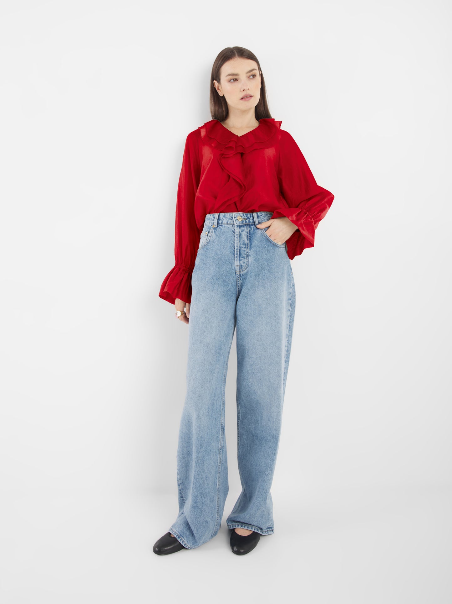 Eine Frau mit glattem braunem Haar und neutralem Gesichtsausdruck modelliert die Ruffle Long Sleeve Red Bluse, gepaart mit hellblauen High-Waisted-Weithalsjeans und schwarzen Schuhen - ein müheloser, nachhaltiger Stil vor einem weißen Hintergrund.