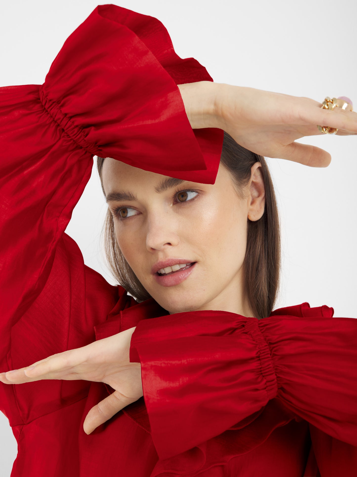 Eine Frau in einer roten Bluse mit voluminösen Rüschenärmeln hebt die Arme, verschränkt sie vor dem Gesicht und blickt zur Seite - ein eleganter Hinweis auf nachhaltige Mode.