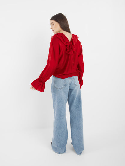 Eine Frau mit langen braunen Haaren trägt das rote Langarm-Oberteil mit Rüschen und eine hellblaue Jeans mit weitem Bein. Sie steht mit dem Gesicht zur Kamera vor einem schlichten weißen Hintergrund.