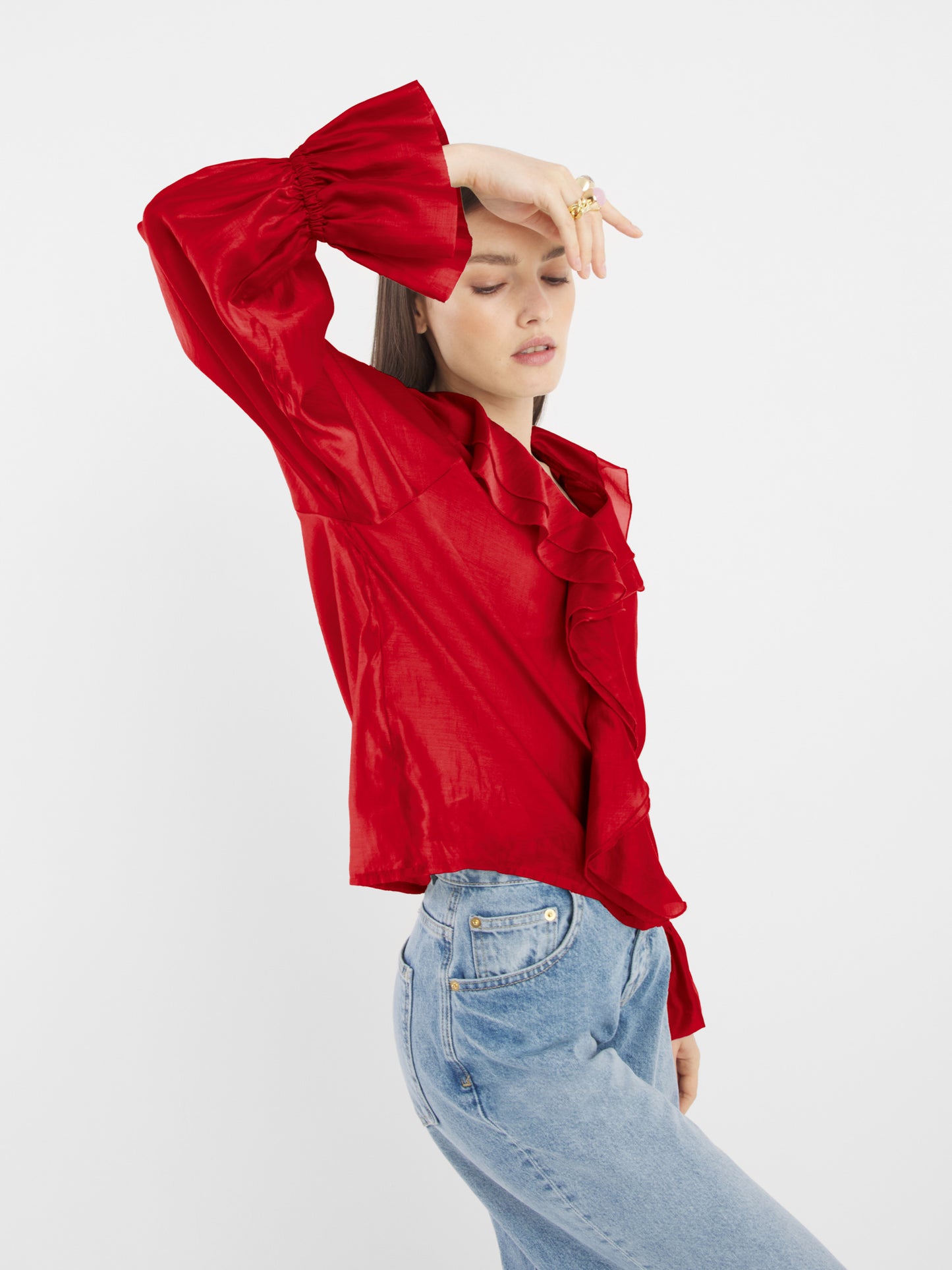 Eine Person posiert seitwärts in einem roten Langarm-Oberteil mit Rüschen und hellblauen Jeans, einen Arm erhoben und die Hand auf der Stirn, vor einem schlichten weißen Hintergrund.