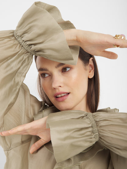 Eine Frau posiert mit erhobenen Armen, die ihr Gesicht betonen, während sie die Ruffle Long Sleeve Taupe Bluse trägt. Das Bild konzentriert sich auf ihren Oberkörper vor einem schlichten Hintergrund. Diese nachhaltige Damenbluse wird in Italien hergestellt.