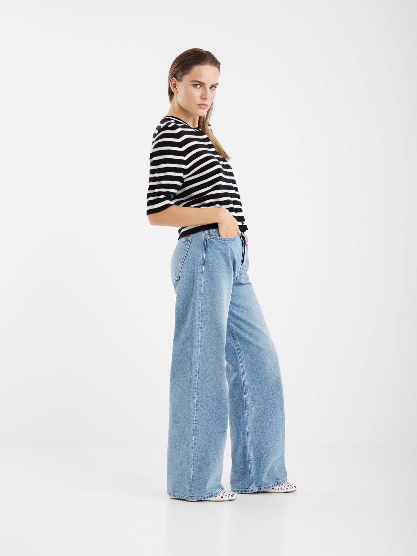 Eine Person posiert in einem Studio vor einem weißen Hintergrund und trägt die Sailor Jacket Short Blue White Stripes und eine hellblaue Jeans mit hoher Taille und weitem Bein, wobei sie eine Hand in der Tasche hat.