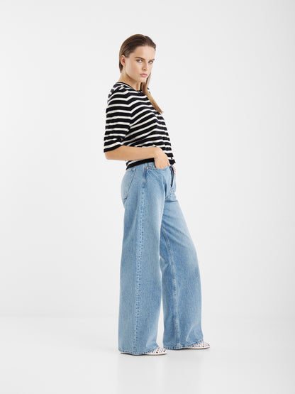 Eine Person posiert in einem Studio vor einem weißen Hintergrund und trägt die Sailor Jacket Short Blue White Stripes und eine hellblaue Jeans mit hoher Taille und weitem Bein, wobei sie eine Hand in der Tasche hat.