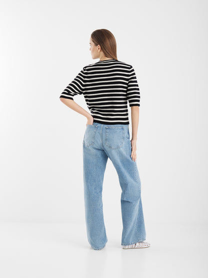 Eine Frau steht mit dem Gesicht zur Wand und trägt die Sailor Jacket Short Blue White Stripes zu hellblauen, weit geschnittenen Jeans. Sie hat glattes braunes Haar und stützt eine Hand auf die Hüfte. Der Hintergrund ist einfarbig weiß.