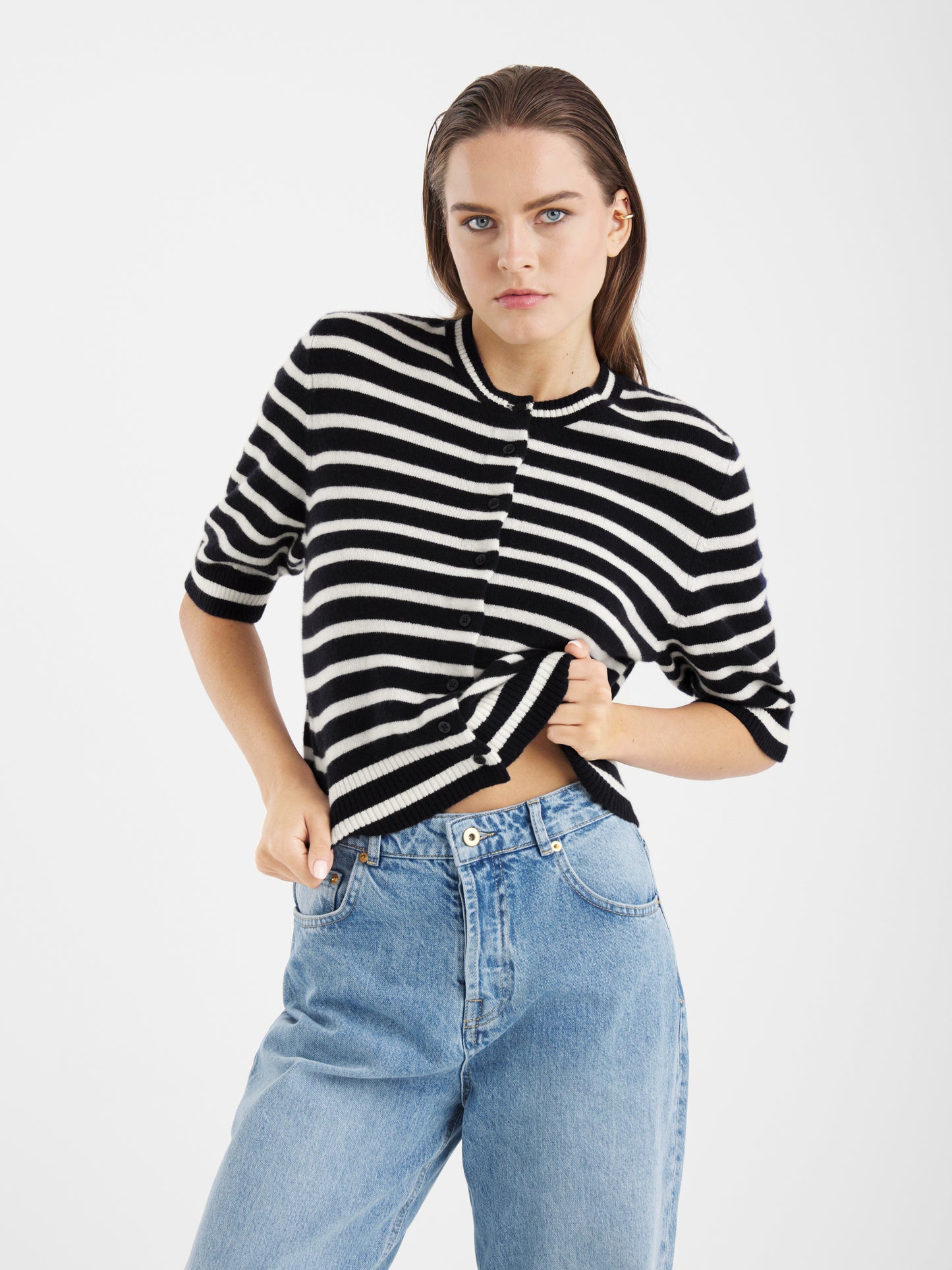 Eine Person mit langen braunen Haaren trägt die Sailor Jacket Short Blue White Stripes und hellblaue Jeans mit hoher Taille, steht vor einem schlichten weißen Hintergrund und blickt selbstbewusst in die Kamera.