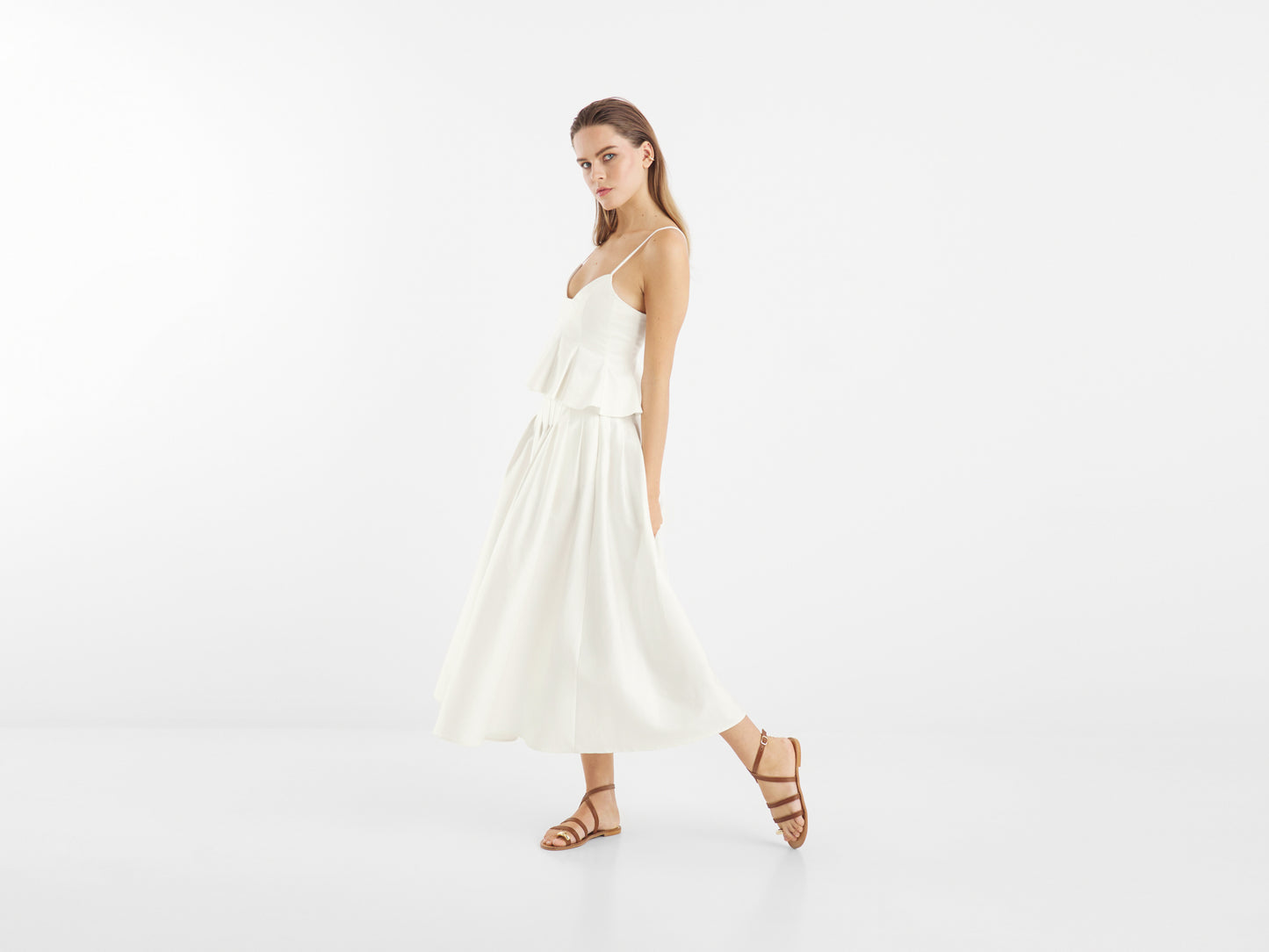 Eine Frau in einem ärmellosen weißen Kleid und braunen Sandalen steht vor einem schlichten Hintergrund, ein Bein leicht angehoben, was den Rock Audrey White hervorhebt - eine elegante Wahl für Sommerrock Damen.