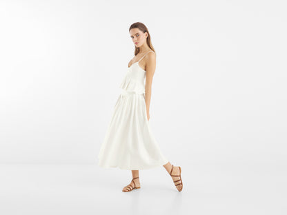 Eine Frau in einem ärmellosen weißen Kleid und braunen Sandalen steht vor einem schlichten Hintergrund, ein Bein leicht angehoben, was den Rock Audrey White hervorhebt - eine elegante Wahl für Sommerrock Damen.