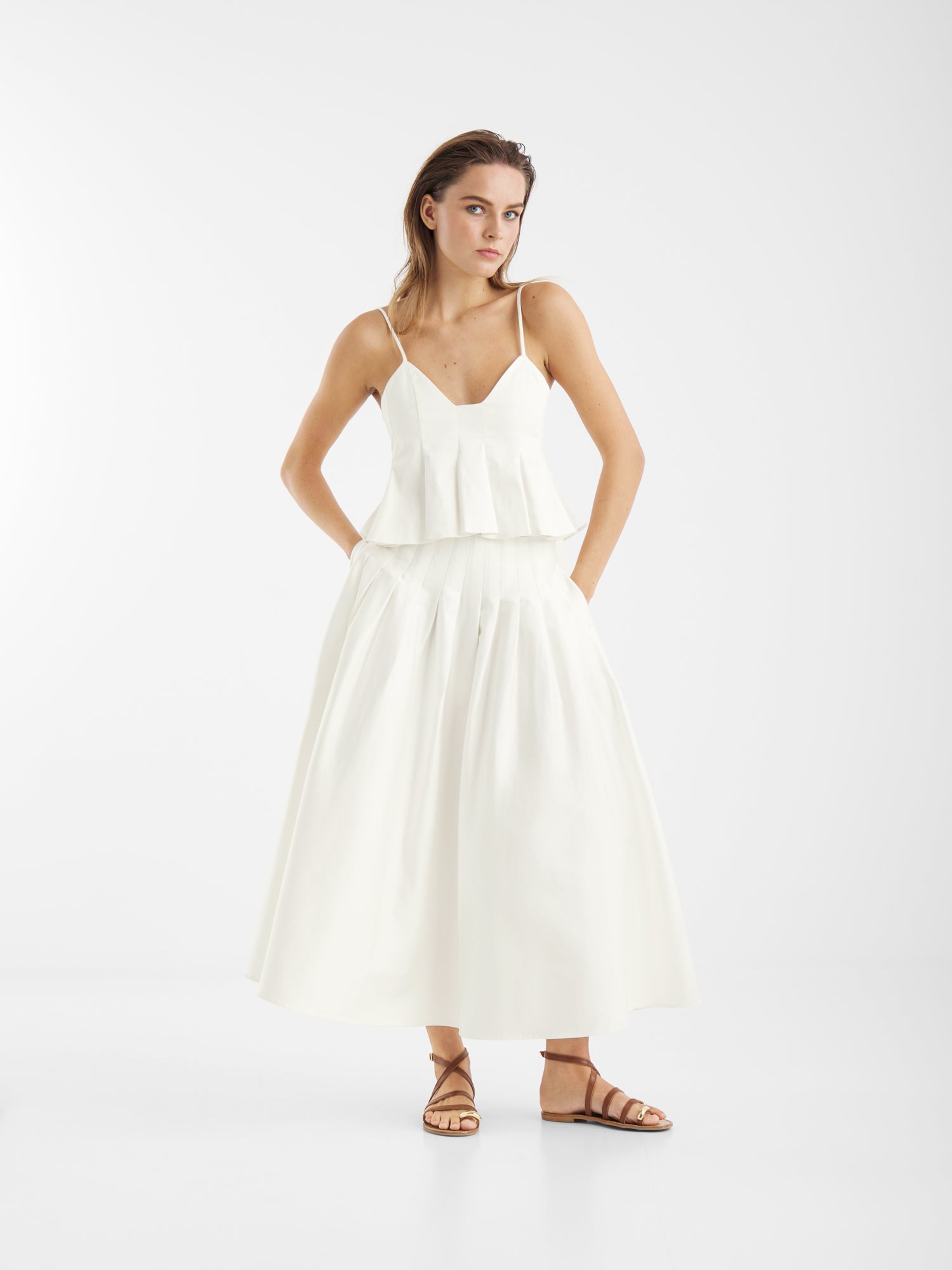 Eine Frau steht vor einem weißen Hintergrund und trägt ein ärmelloses weißes Oberteil mit Schößchen, den Rock Audrey White aus Baumwolle als Sommerrock für Frauen und braune flache Sandalen. Ihr Haar ist glatt und ihre Hände sind in ihren Taschen.