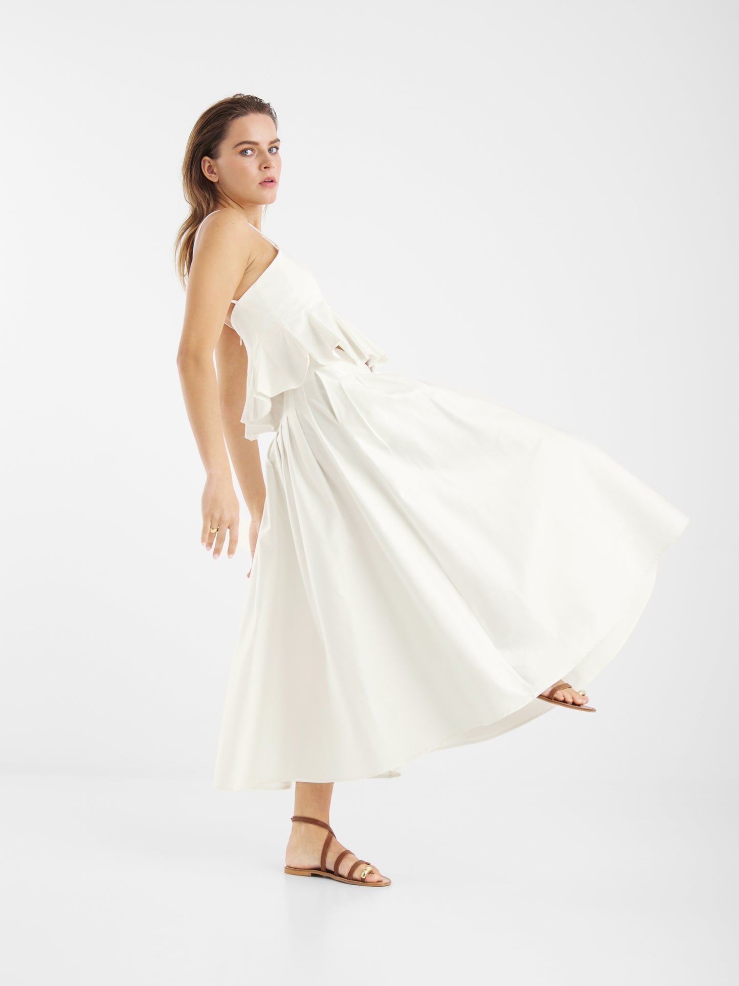 Eine Frau in einem fließenden, ärmellosen weißen Kleid und braunen Sandalen steht seitlich und strahlt mühelose Anmut aus, während sie den Rock Audrey White for Damen vor einem schlichten weißen Hintergrund präsentiert.