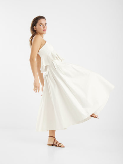 Eine Frau in einem fließenden, ärmellosen weißen Kleid und braunen Sandalen steht seitlich und strahlt mühelose Anmut aus, während sie den Rock Audrey White for Damen vor einem schlichten weißen Hintergrund präsentiert.