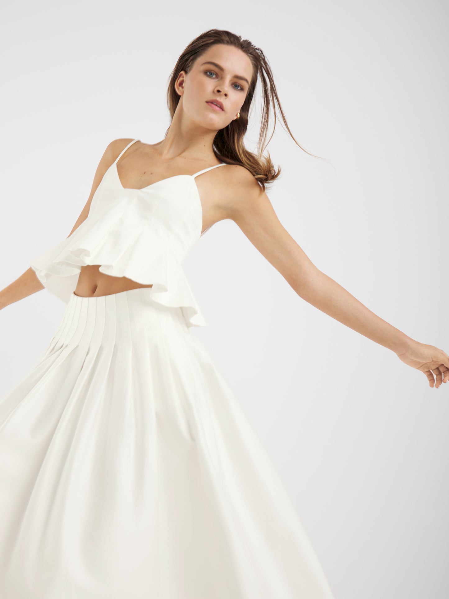Eine Frau wirbelt anmutig vor einem schlichten, hellen Hintergrund. Sie trägt ein ärmelloses weißes Crop-Top mit Rüschen und den zeitlos stilvollen Rock Audrey White, der den eleganten Charme dieses Sommerrocks Damen unterstreicht.