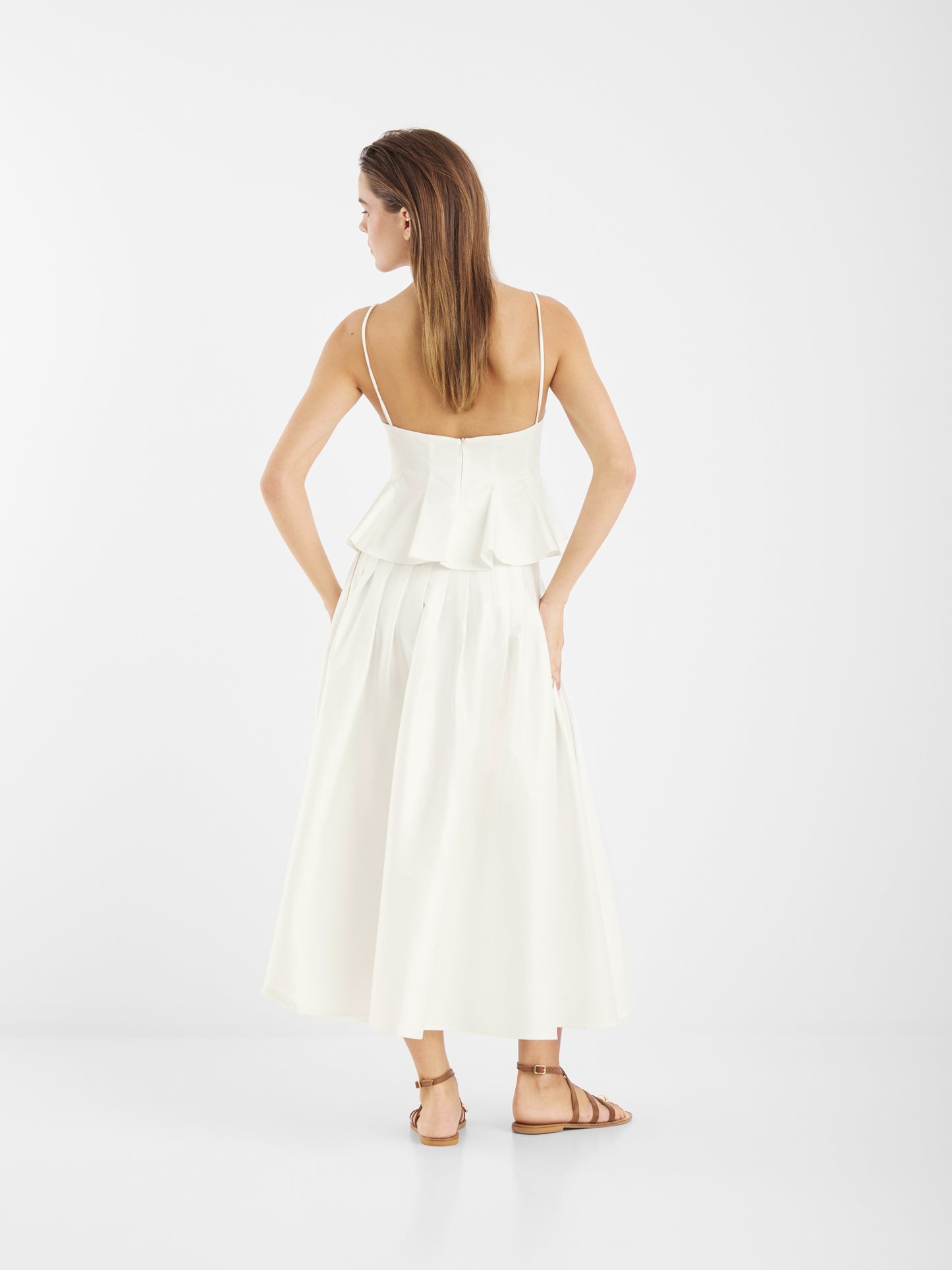Eine Frau mit glattem braunem Haar und flachen braunen Sandalen blickt nach vorne. Sie trägt ein ärmelloses weißes Rüschenoberteil und den knöchellangen Baumwollrock Audrey White vor einem schlichten weißen Hintergrund.