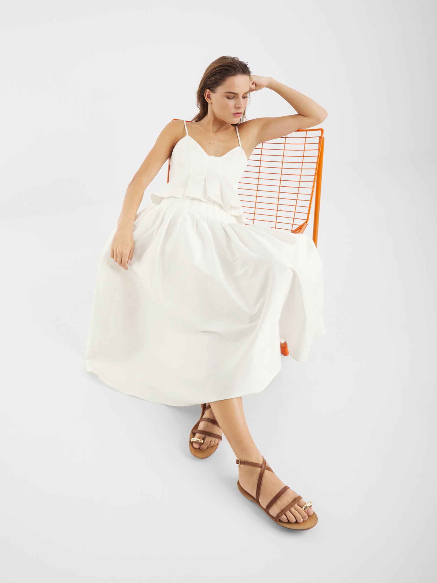 Eine Frau, die das Top Audrey White trägt, sitzt entspannt auf einem orangefarbenen Drahtstuhl, den Kopf auf eine Hand gestützt. Braune Riemchensandalen und ein schlichter weißer Hintergrund vervollständigen ihren mühelosen Sommer-Essential-Look.