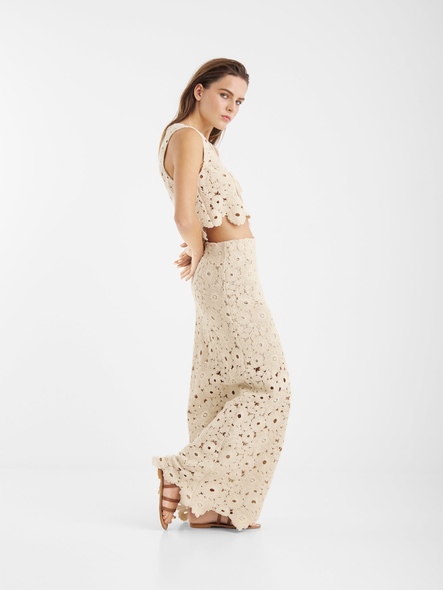 Eine Frau mit langen Haaren posiert seitlich vor einem weißen Hintergrund. Sie trägt den Skirt Ibiza Beige - ein beigefarbenes, ärmelloses Crop-Top aus Häkelarbeit mit einem passenden langen Rock mit Blumenmuster - und dazu flache Sandalen.