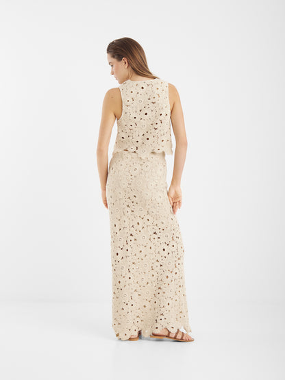 Eine Frau blickt weg und trägt die Vest Ibiza - ein ärmelloses, cremefarbenes, knöchellanges Häkelkleid mit Blumenmuster und Sandalen - vor einem schlichten weißen Hintergrund.