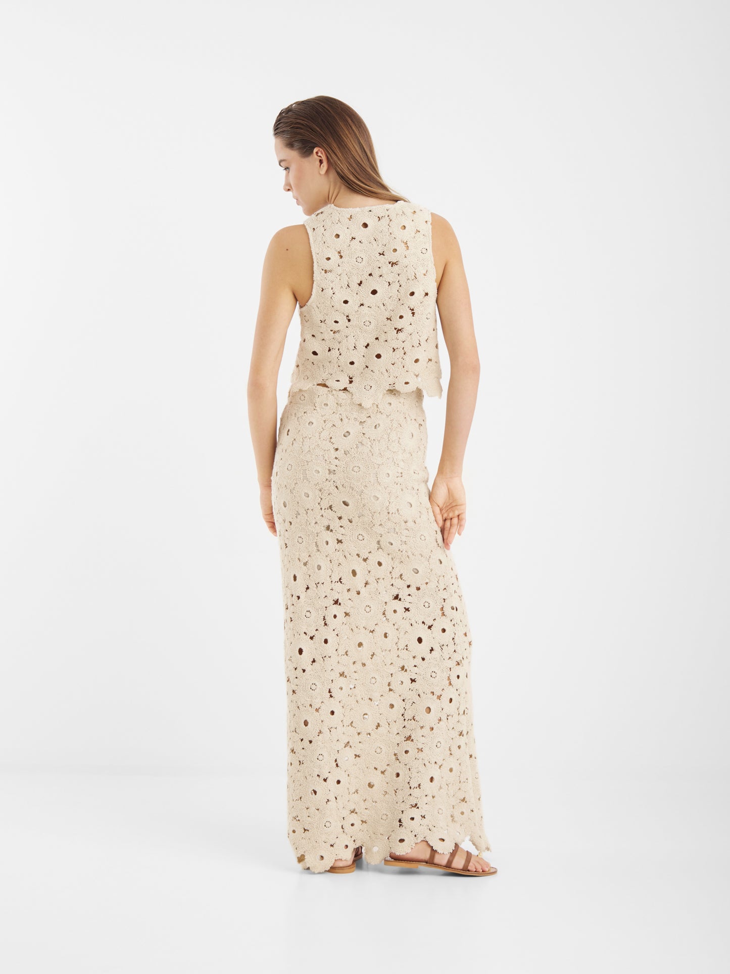 Eine Frau steht mit dem Rock Ibiza Beige - einem ärmellosen, beigefarbenen, häkelgemusterten Oberteil und einem dazu passenden langen Rock mit Sandalen - vor einem einfarbig weißen Hintergrund.