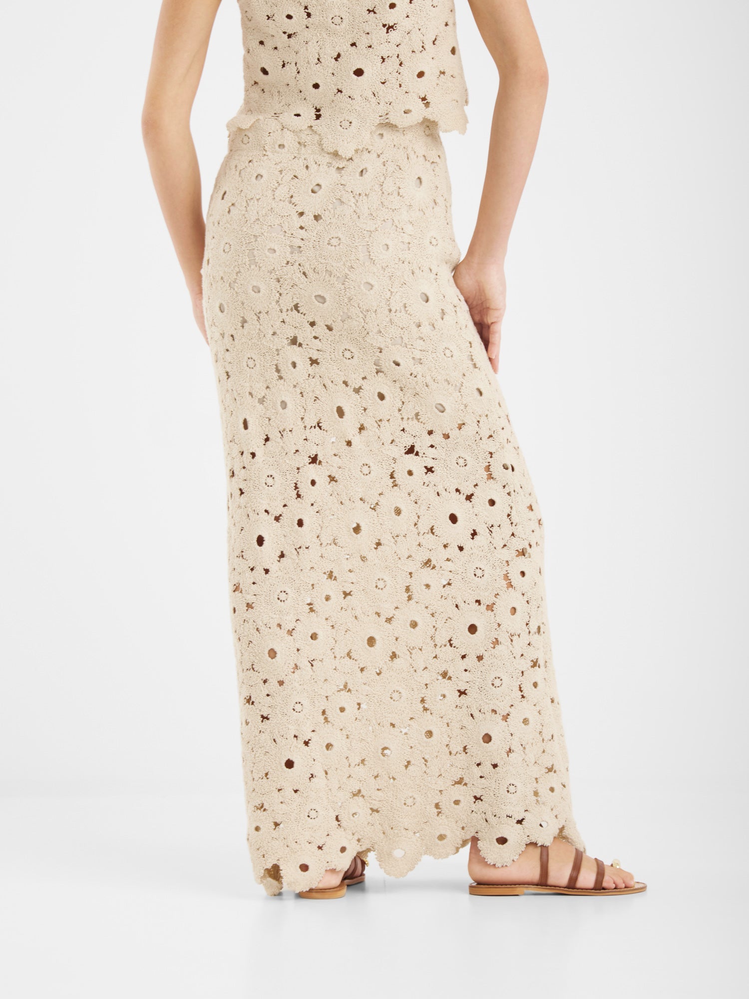 Eine Person trägt den cremefarbenen, knöchellangen Vest Ibiza-Häkelrock mit überschnittenem Saum und das passende Häkeloberteil, gepaart mit flachen braunen Sandalen. Sie steht seitlich vor einem schlichten weißen Hintergrund.