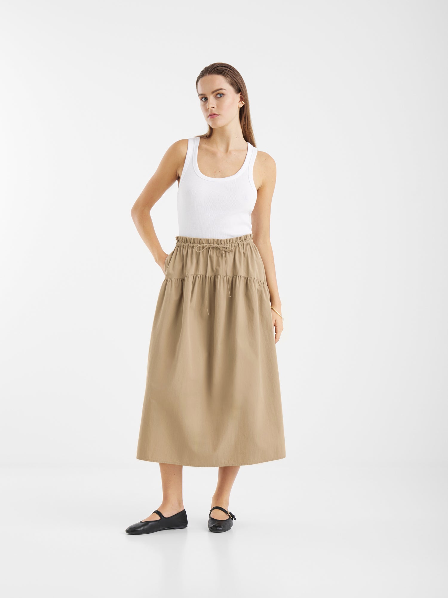 Eine Frau steht vor einem weißen Hintergrund und trägt ein weißes Tanktop, den Rock Palma Beige mit elastischem Bund und schwarze Flats. Sie sieht neutral aus und hat die Hände in den Rocktaschen.