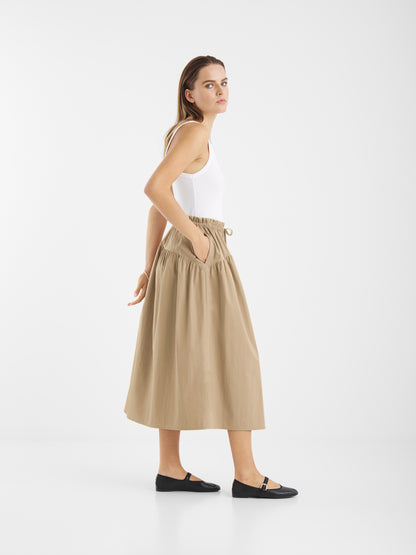 Eine Frau steht seitlich vor einem weißen Hintergrund. Sie trägt ein weißes ärmelloses Oberteil, den Rock Palma Beige mit Taschen und schwarze Flats. Sie blickt mit neutralem Blick über ihre Schulter.