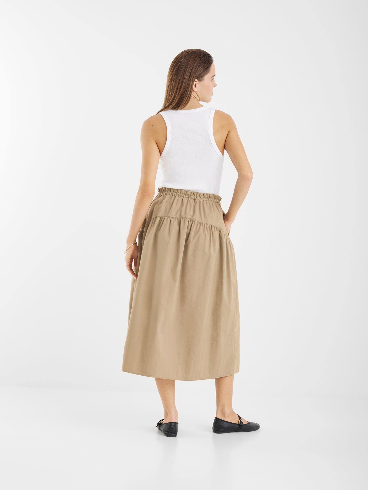 Eine Frau steht mit dem Gesicht zur Wand und trägt ein weißes ärmelloses Oberteil, den Rock Palma Beige (hochtaillierter Midirock aus Baumwolle) und schwarze flache Schuhe vor einem schlichten weißen Hintergrund.