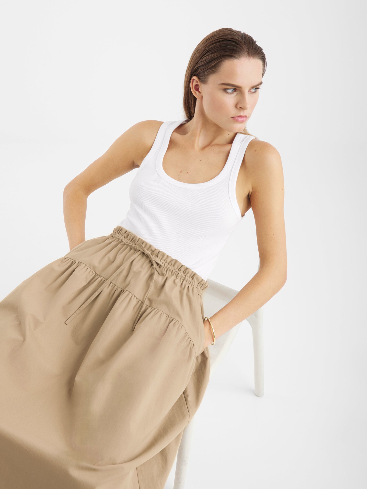Eine Frau mit glattem braunem Haar trägt ein weißes Tanktop und den Rock Palma Beige. Sie sitzt auf einem weißen Stuhl und schaut mit den Händen in den Rocktaschen vor einem schlichten weißen Hintergrund zur Seite.