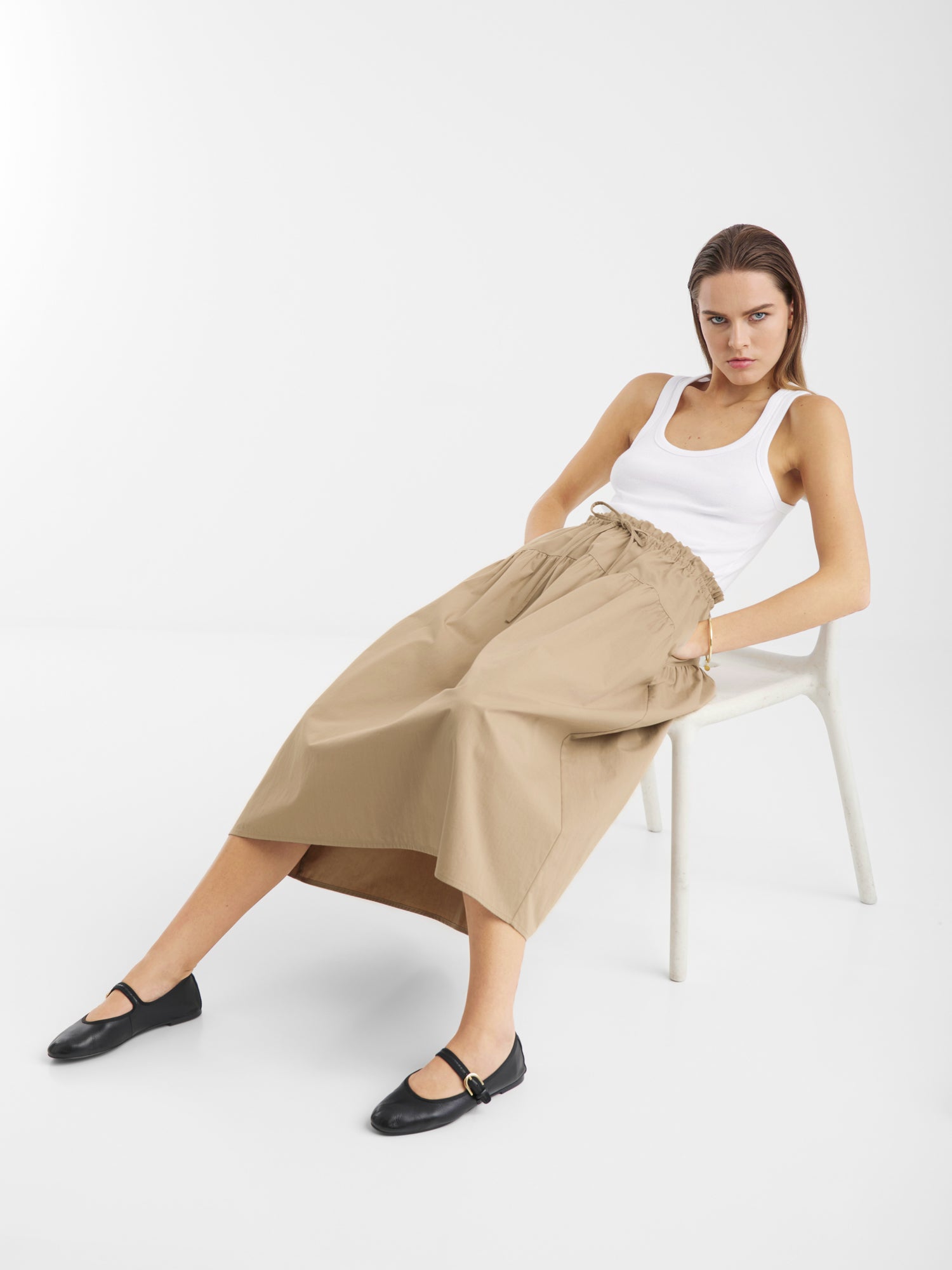 Eine Frau sitzt auf einem weißen Stuhl vor einem schlichten Hintergrund und trägt ein weißes Tanktop, den Rock Palma Beige mit elastischem Bund und schwarze Mary-Jane-Schuhe. Sie sieht entspannt aus, hat die Beine gespreizt und die Arme auf den Knien abgestützt.