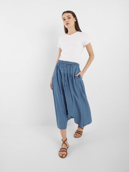 Eine Frau steht vor einem weißen Hintergrund und trägt ein schlichtes weißes T-Shirt, den weiten blauen Rock Palma Denim mit Taschen und braune Riemchensandalen. Sie hat glattes braunes Haar und einen neutralen Gesichtsausdruck.