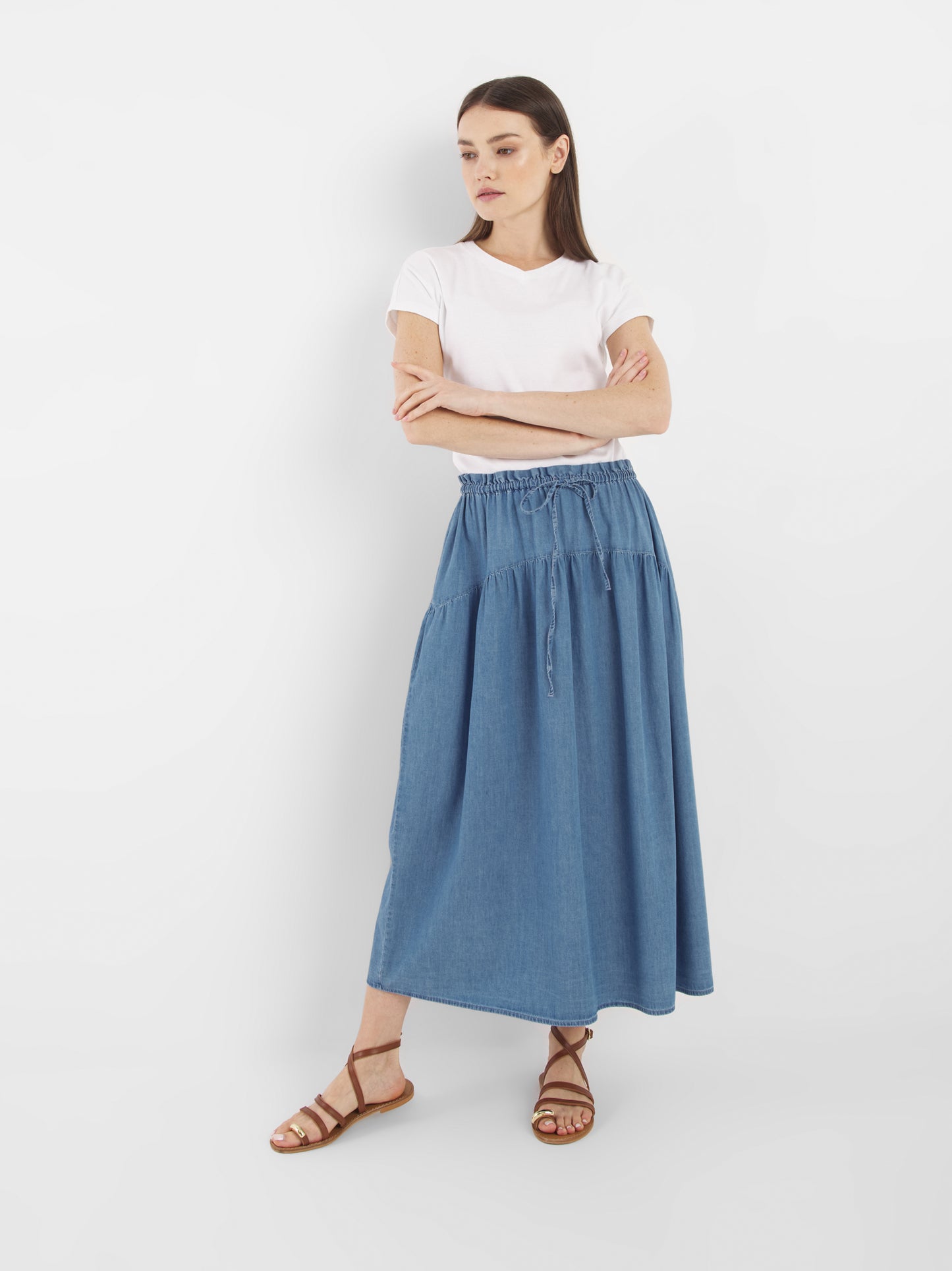 Eine Frau steht vor einem weißen Hintergrund, trägt ein weißes T-Shirt, den Rock Palma Denim in blauem, knöchellangem Stil und braune Riemchensandalen, die Arme verschränkt und schaut zur Seite.
