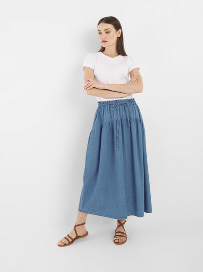 Eine Frau steht vor einem weißen Hintergrund, trägt ein weißes T-Shirt, den Rock Palma Denim in blauem, knöchellangem Stil und braune Riemchensandalen, die Arme verschränkt und schaut zur Seite.