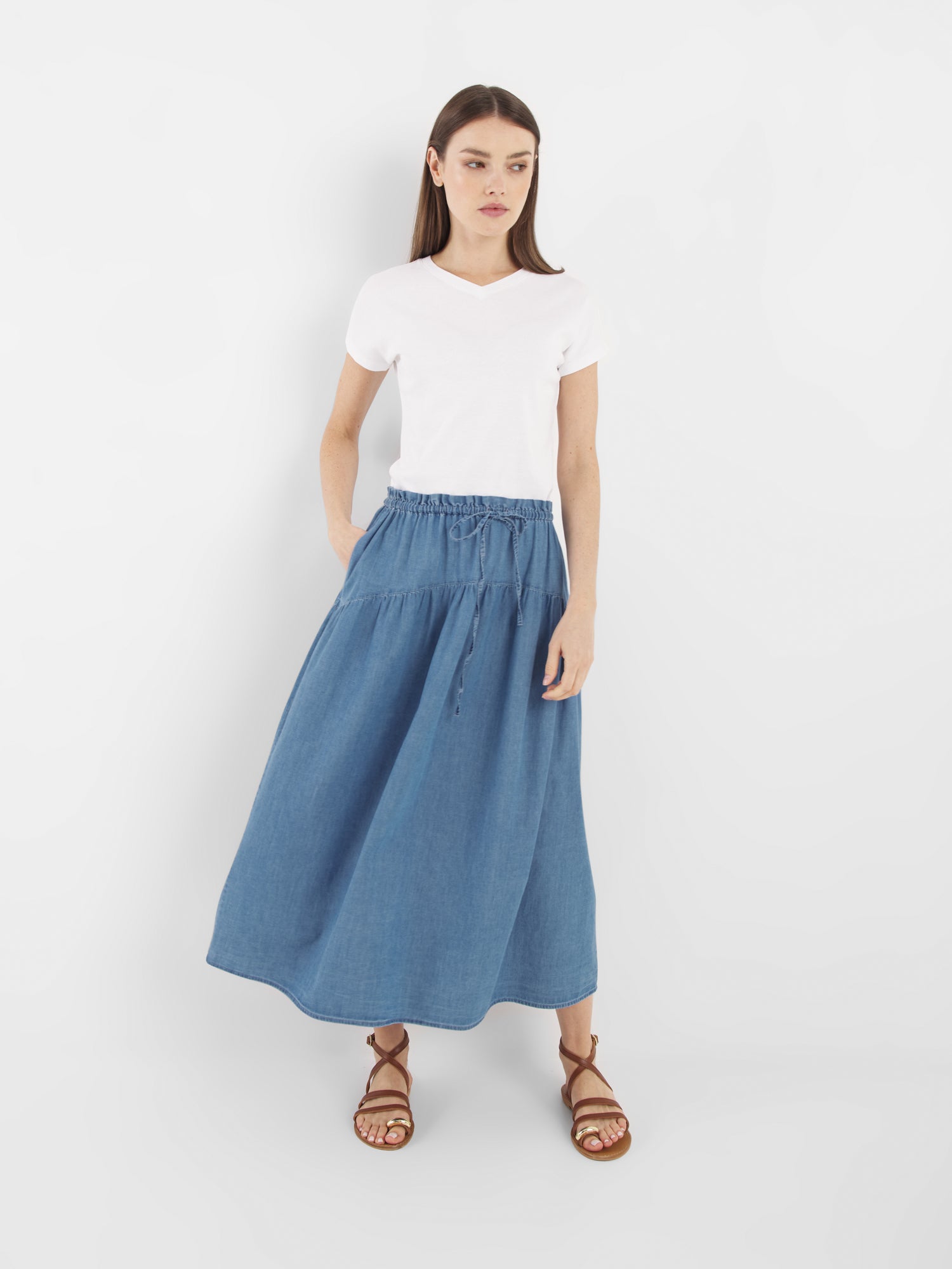 Eine Frau mit glattem braunem Haar und neutralem Gesichtsausdruck steht vor einem weißen Hintergrund. Sie trägt ein schlichtes weißes T-Shirt, den Skirt Palma Denim - einen blauen Jeans-Midirock mit Kordelzug in der Taille - und braune Riemchensandalen.