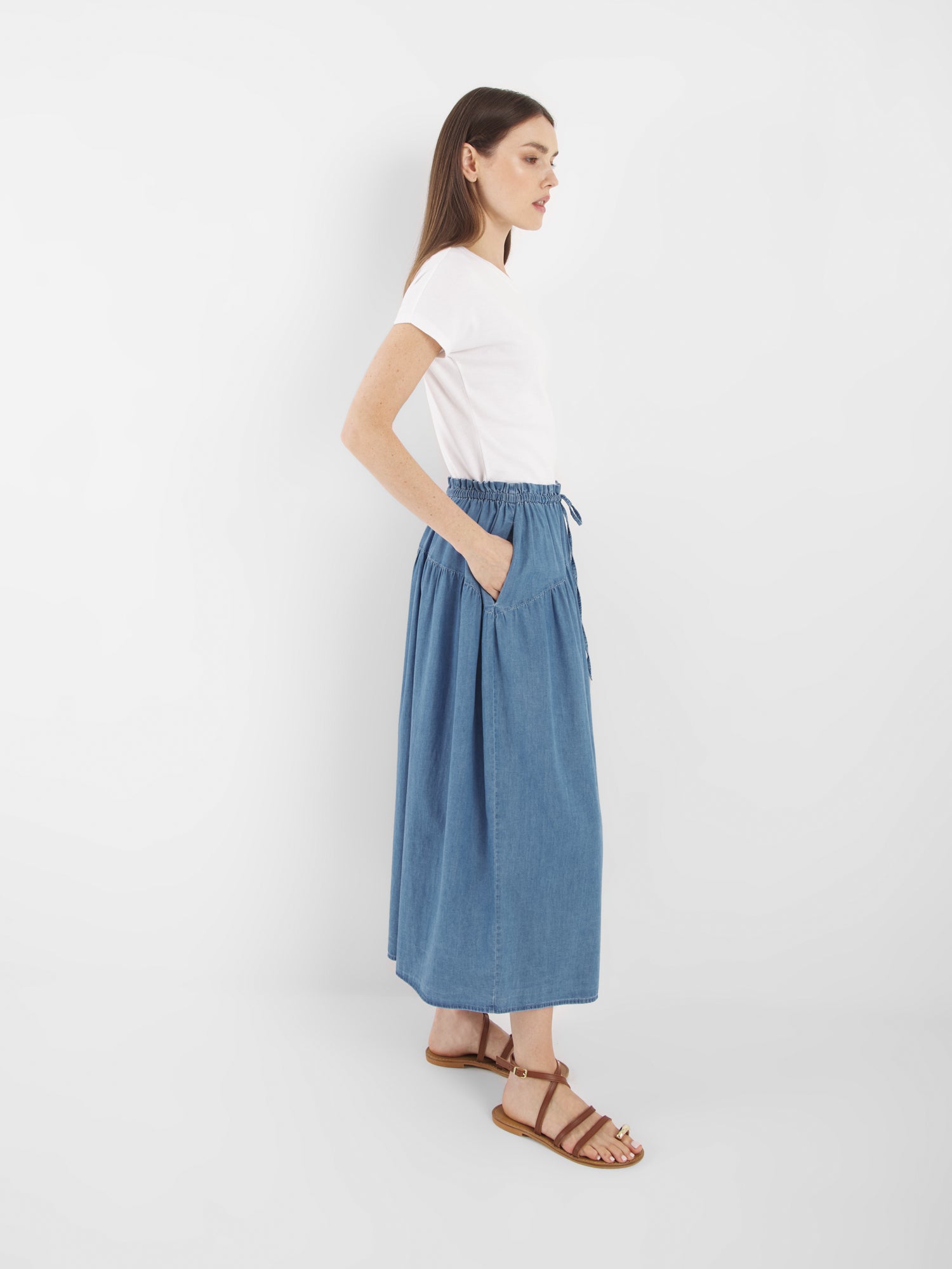 Eine Frau steht seitlich vor einem weißen Hintergrund und trägt ein weißes T-Shirt, braune Sandalen und den Rock Palma Denim. Ihr glattes braunes Haar und die lässigen Hände in den Rocktaschen vervollständigen den lässigen Look.