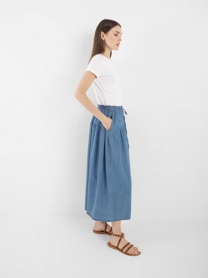 Eine Frau steht seitlich vor einem weißen Hintergrund und trägt ein weißes T-Shirt, braune Sandalen und den Rock Palma Denim. Ihr glattes braunes Haar und die lässigen Hände in den Rocktaschen vervollständigen den lässigen Look.