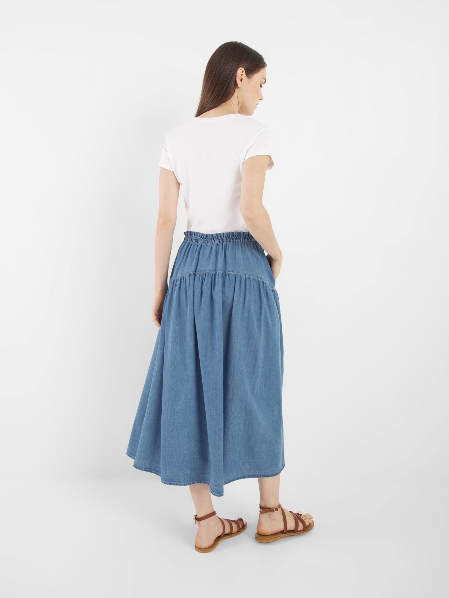 Eine Frau steht mit dem Gesicht zur Wand und trägt ein weißes, kurzärmeliges T-Shirt, den Rock Palma Denim mit geraffter Taille und braune Sandalen vor einem schlichten weißen Hintergrund.