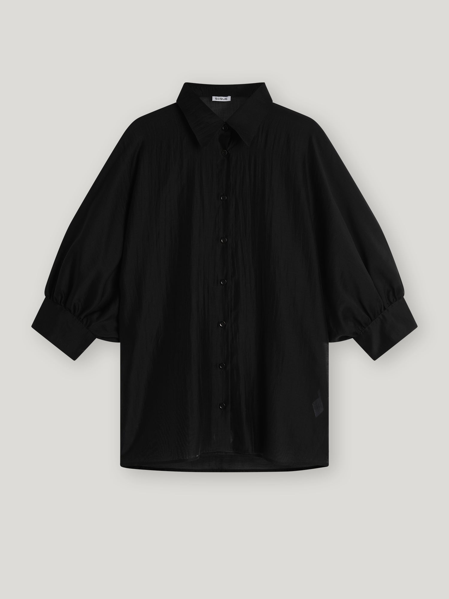 Die Bluse Antonia Short Sleeve Black ist eine locker sitzende schwarze Bluse mit Kragen, übergroßem Schnitt und weiten Dreiviertel-Ärmeln, die an den Manschetten gerafft sind, auf einem hellgrauen Hintergrund.