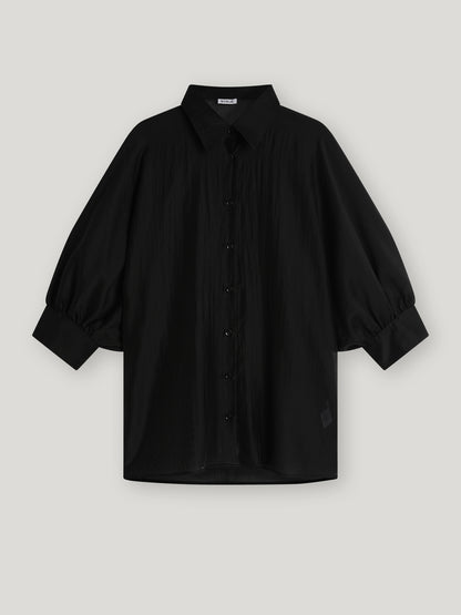 Die Bluse Antonia Short Sleeve Black ist eine locker sitzende schwarze Bluse mit Kragen, übergroßem Schnitt und weiten Dreiviertel-Ärmeln, die an den Manschetten gerafft sind, auf einem hellgrauen Hintergrund.