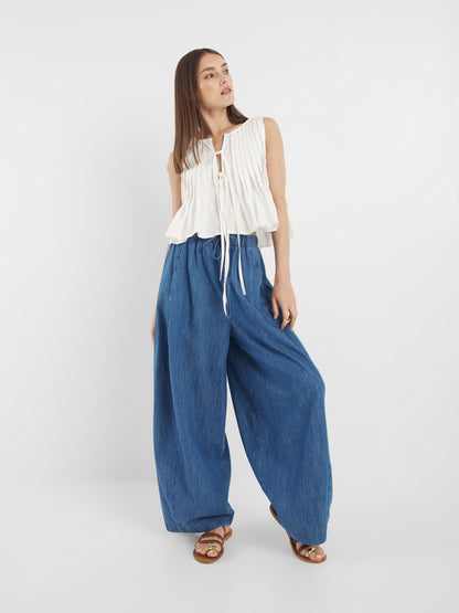 Eine Frau posiert vor einem weißen Hintergrund und trägt das Top Cleo Weiss, ein ärmelloses weißes Oberteil mit Bindebändern auf der Vorderseite, gepaart mit einer weit geschnittenen blauen Jeanshose. Sie hat glattes Haar und trägt braune Sandalen.