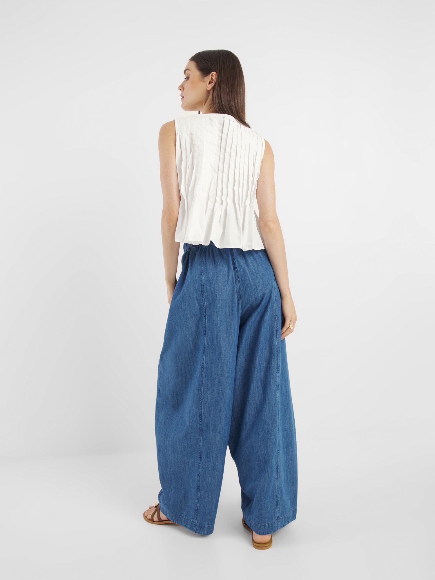 Eine Frau blickt von sich weg und trägt das Top Cleo Weiss - ein ärmelloses weißes Oberteil mit plissiertem Rücken - zusammen mit einer blauen Jeanshose mit weitem Bein und braunen Sandalen vor einem schlichten weißen Hintergrund.